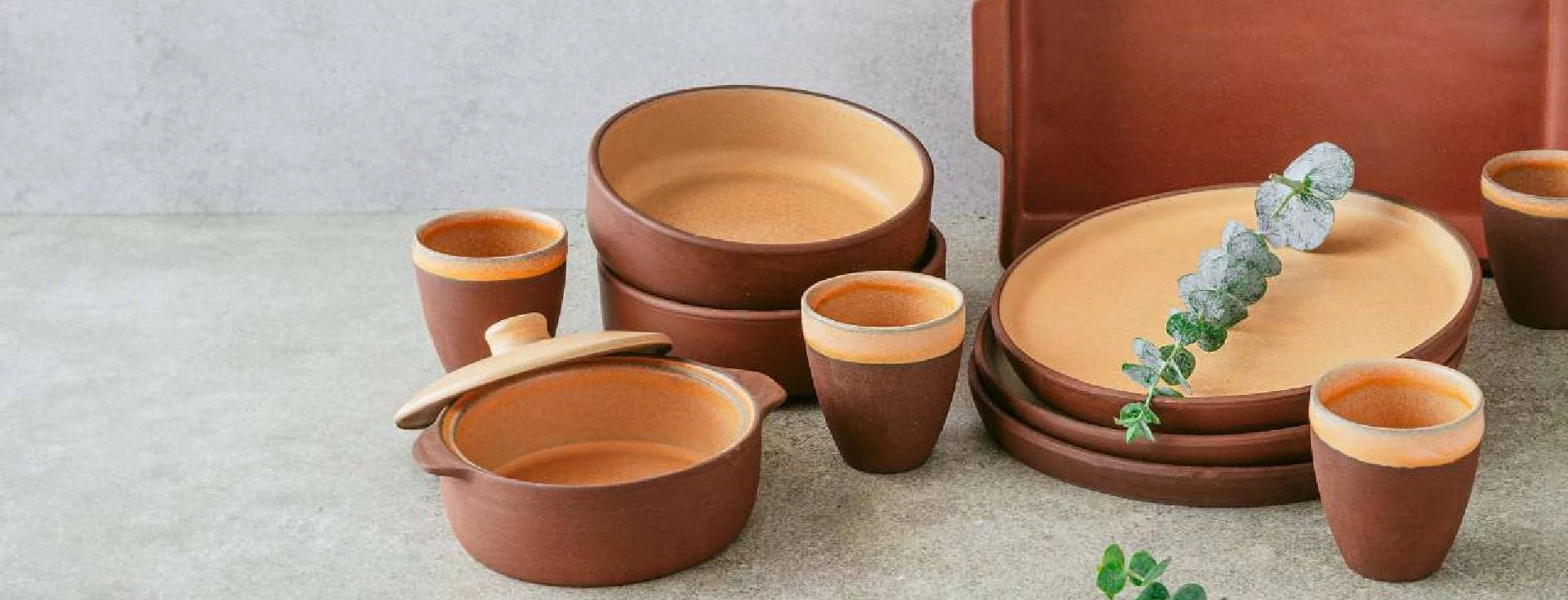 Earthenware dining set with plates, bowls, and cups in rich terracotta. A sprig of eucalyptus adds a touch of green to the rustic display.